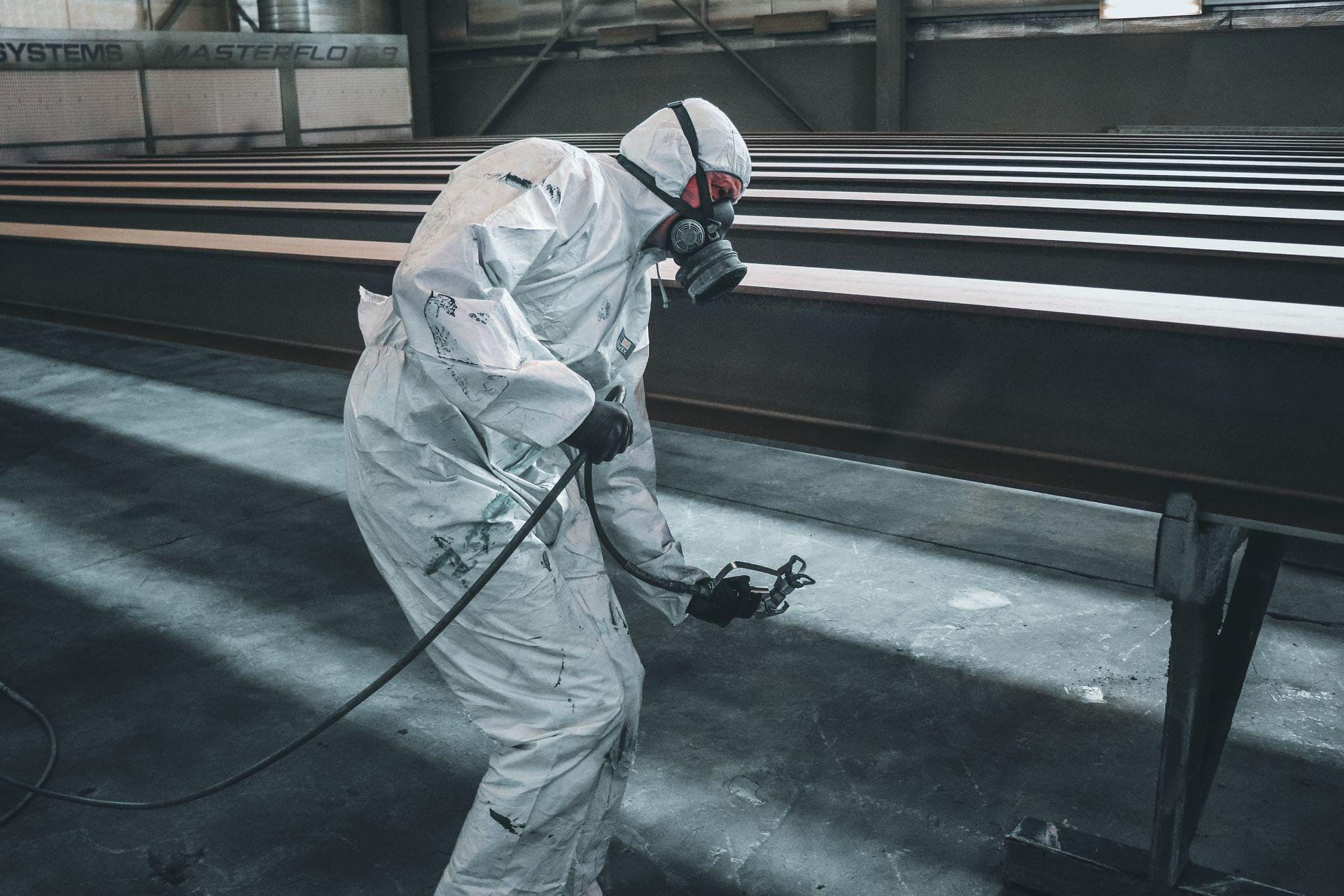 Worker in protective gear spray painting metal beams inside a factory setting.
