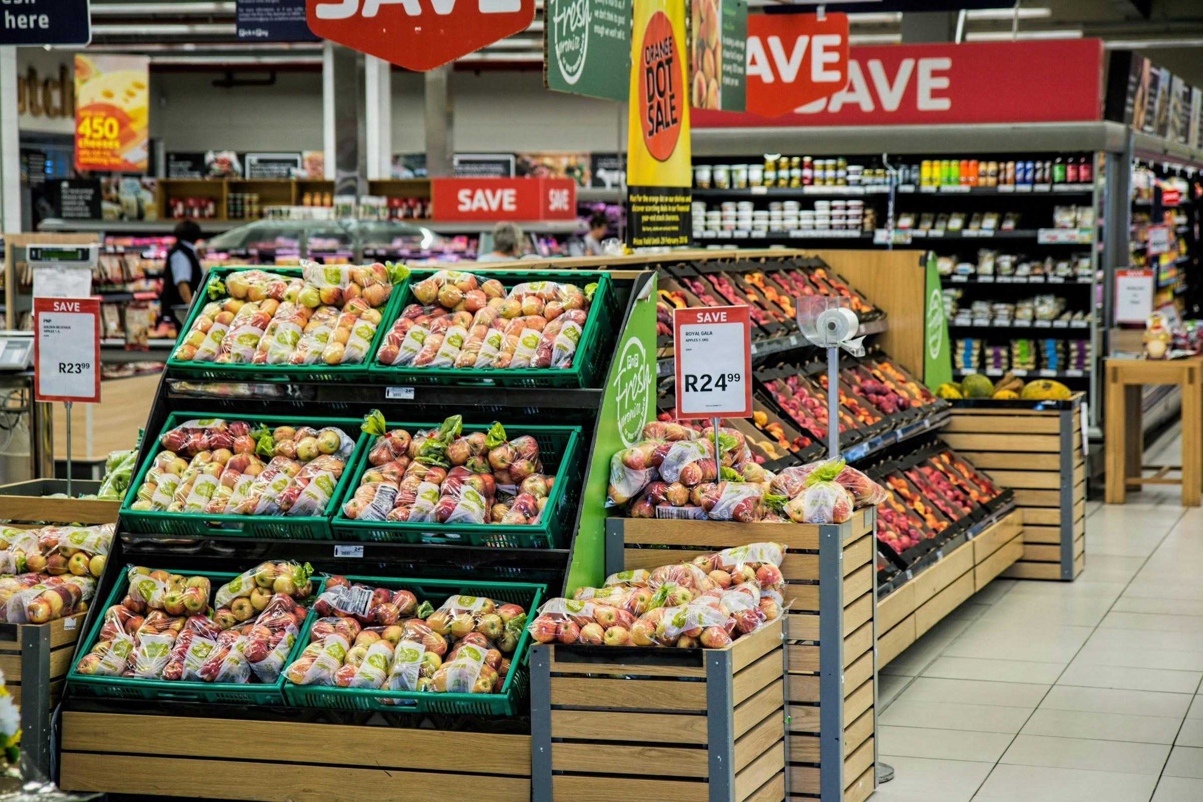 Produce section in a supermarket with bags of apples displayed in green crates and wooden bins